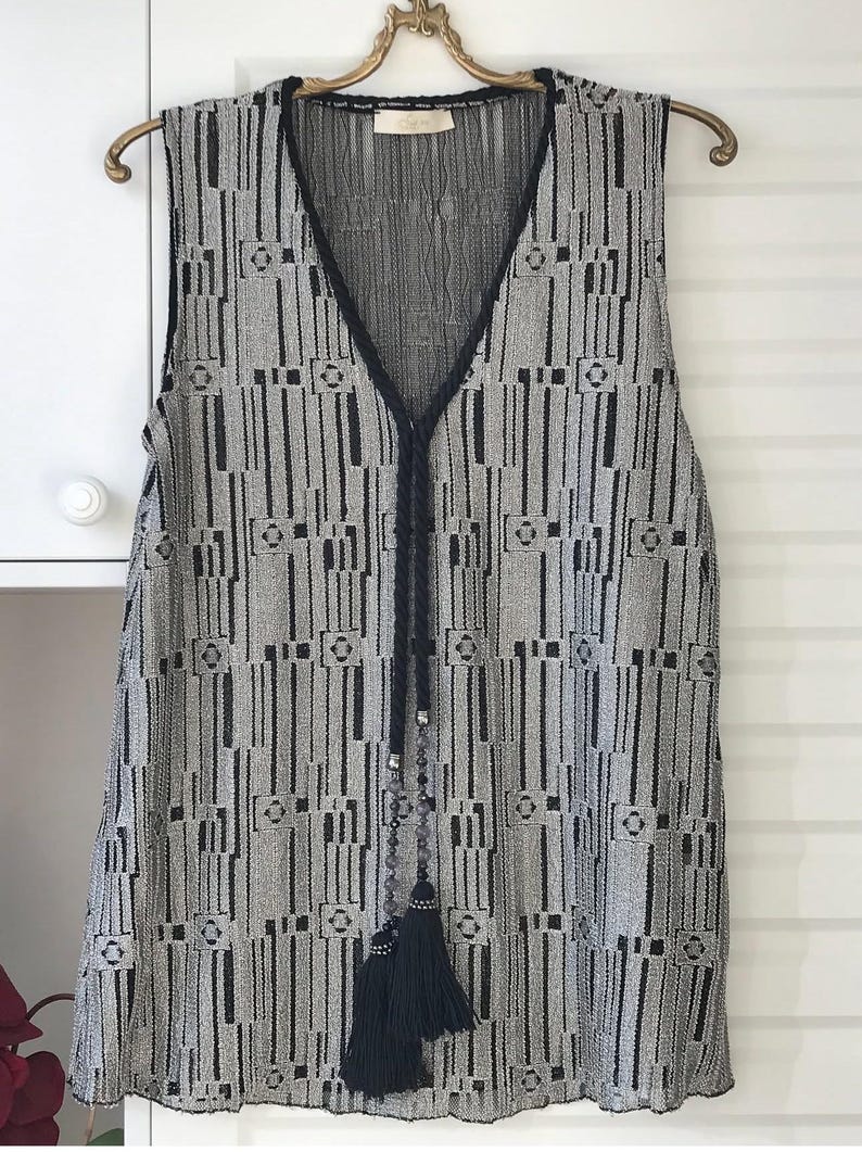 May include: Sleeveless vest featuring a geometric design in black and grey. The vest has a V-neckline trimmed with black fabric and a black braided tie with beaded accents and a tassel. The garment is suspended on a gold-coloured hanger.