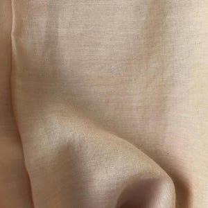 May include: Close-up of a peach-colored fabric with a subtle, textured weave. The material is draped, creating soft folds and shadows. The fabric has a slight sheen, suggesting a delicate or luxurious textile.