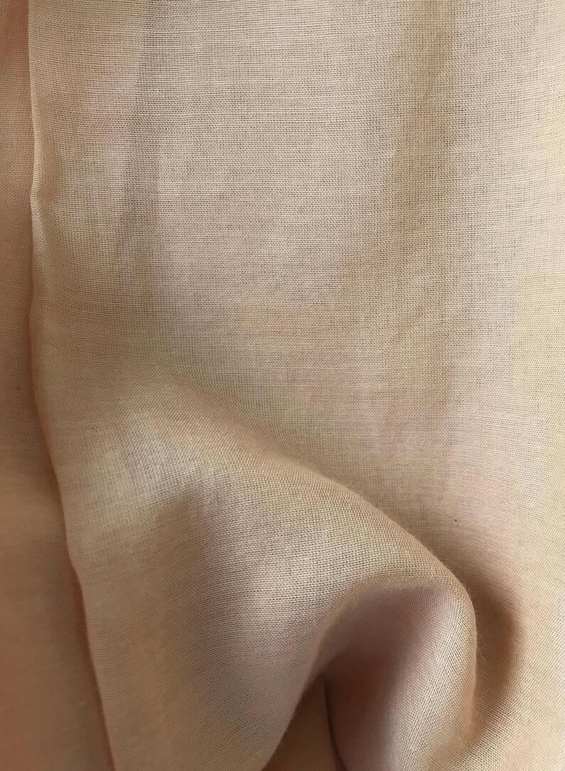 May include: Close-up of a peach-colored fabric with a subtle, textured weave. The material is draped, creating soft folds and shadows. The fabric has a slight sheen, suggesting a delicate or luxurious textile.