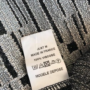 May include: Close-up of a textured fabric with a pattern of vertical black stripes on a silver background. A white tag reads "JUST M MADE IN FRANCE 100% VISCOSE MODELE DEPOSE".