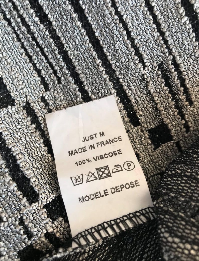May include: Close-up of a textured fabric with a pattern of vertical black stripes on a silver background. A white tag reads "JUST M MADE IN FRANCE 100% VISCOSE MODELE DEPOSE".