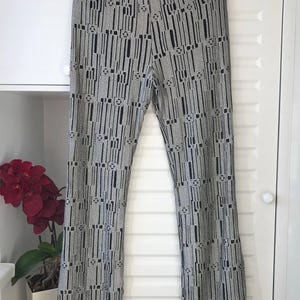 May include: Patterned trousers in black and silver, featuring a high waist with a black sequined band. The design includes vertical black lines and geometric shapes on a silver backdrop. The trousers are displayed against a white door.