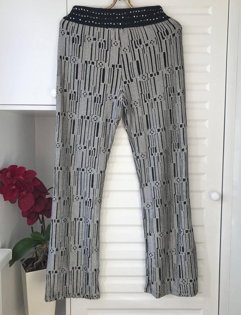 May include: Patterned trousers in black and silver, featuring a high waist with a black sequined band. The design includes vertical black lines and geometric shapes on a silver backdrop. The trousers are displayed against a white door.