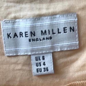 May include: A close-up of a clothing tag with the brand name "KAREN MILLEN ENGLAND" in black letters. Below the brand name, a smaller tag indicates sizes: UK 8, US 4, and EU 36. The background is a light peach color.