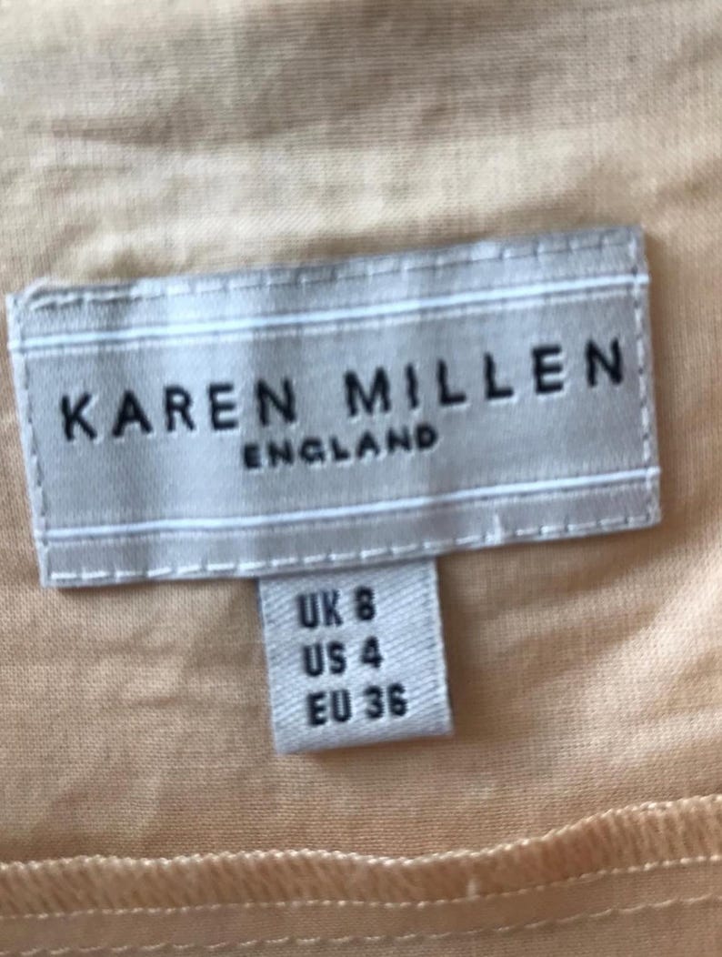May include: A close-up of a clothing tag with the brand name "KAREN MILLEN ENGLAND" in black letters. Below the brand name, a smaller tag indicates sizes: UK 8, US 4, and EU 36. The background is a light peach color.