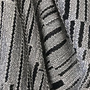 May include: Close-up of a textured fabric with a woven pattern. The fabric features a mix of grey and black threads, creating a striped design. The texture appears slightly raised, adding depth to the material. The fabric is folded, showing the interplay of light and shadow.