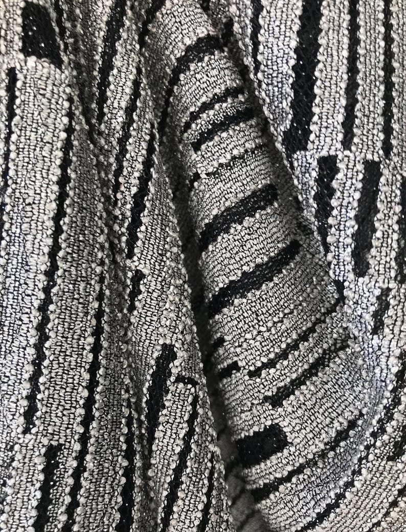 May include: Close-up of a textured fabric with a woven pattern. The fabric features a mix of grey and black threads, creating a striped design. The texture appears slightly raised, adding depth to the material. The fabric is folded, showing the interplay of light and shadow.