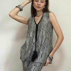 May include: A silver and black patterned sleeveless top and matching trousers. The outfit features a V-neck and a black tassel. The model is wearing silver bracelets and earrings. The outfit is a two-piece set.