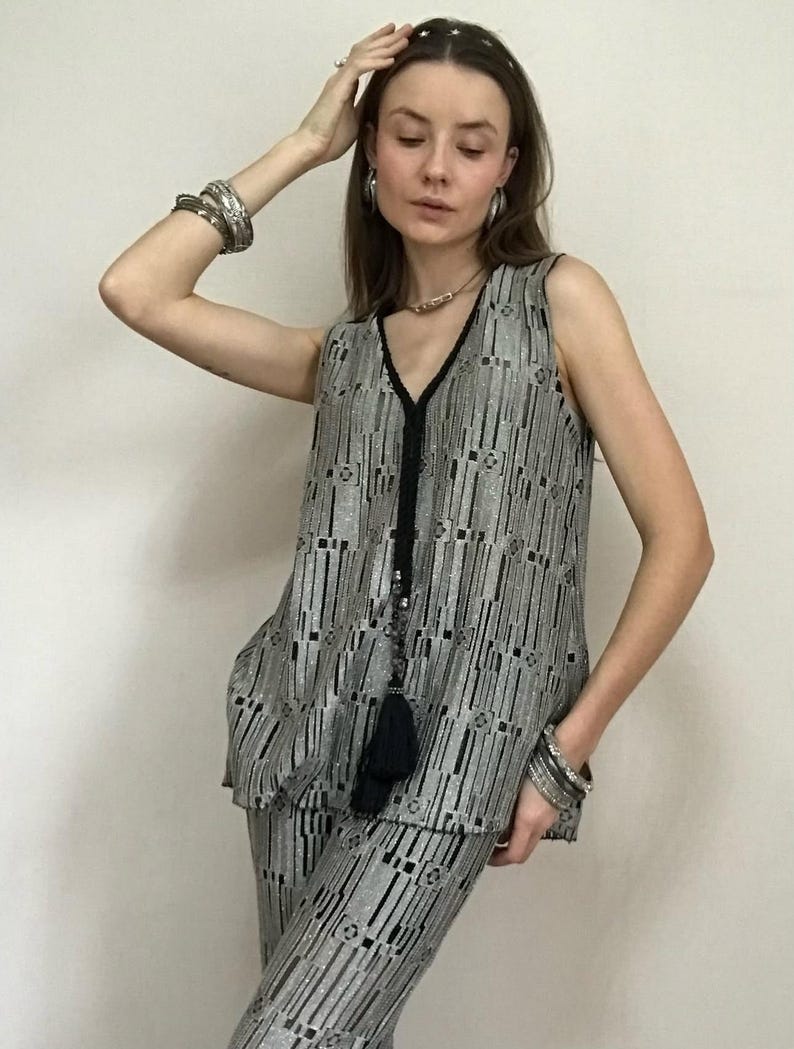 May include: A silver and black patterned sleeveless top and matching trousers. The outfit features a V-neck and a black tassel. The model is wearing silver bracelets and earrings. The outfit is a two-piece set.