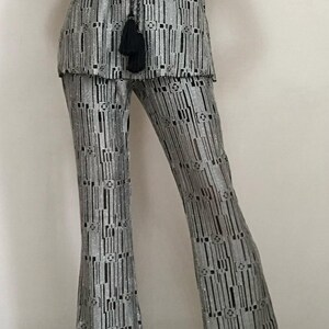May include: Silver and black patterned two-piece outfit. The top is sleeveless, and the trousers are flared. The fabric has a vertical stripe design with geometric accents. A black tassel necklace is visible.