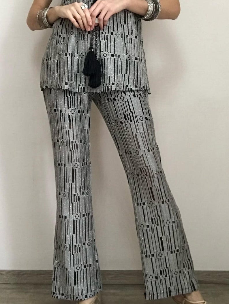 May include: Silver and black patterned two-piece outfit. The top is sleeveless, and the trousers are flared. The fabric has a vertical stripe design with geometric accents. A black tassel necklace is visible.