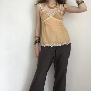 May include: A tan camisole with lace trim and brown pants. The camisole has a delicate lace trim along the neckline and hem. The pants are a dark brown color and have a wide-leg design. The outfit is accessorized with gold jewelry and bracelets.