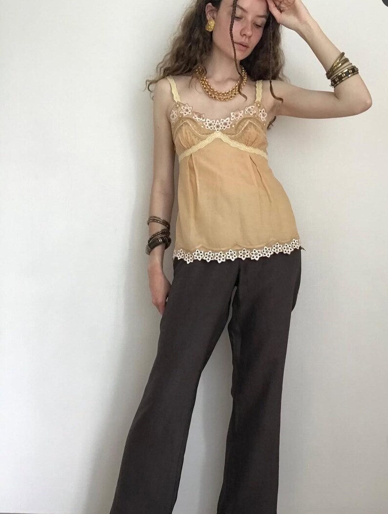 May include: A tan camisole with lace trim and brown pants. The camisole has a delicate lace trim along the neckline and hem. The pants are a dark brown color and have a wide-leg design. The outfit is accessorized with gold jewelry and bracelets.