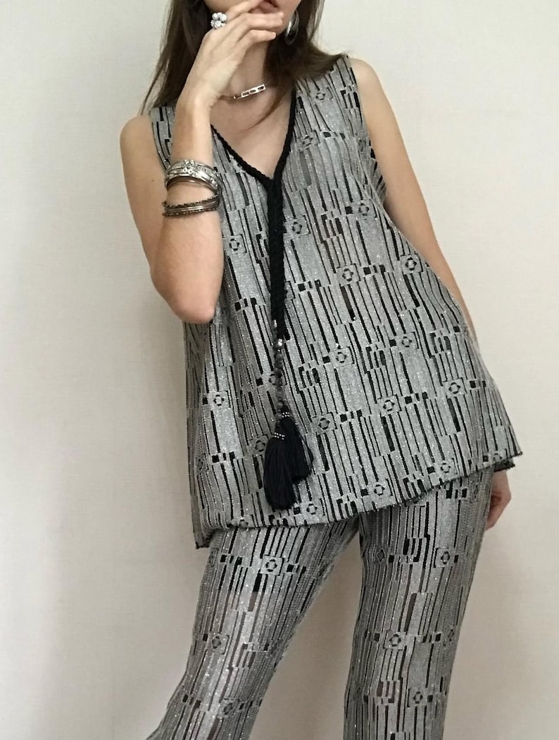 May include: Silver sleeveless top and matching trousers set. The outfit has a vertical striped pattern with black accents. A black tassel hangs from the V-neckline of the top. The fabric has a shimmering, textured appearance, creating a glamorous look.