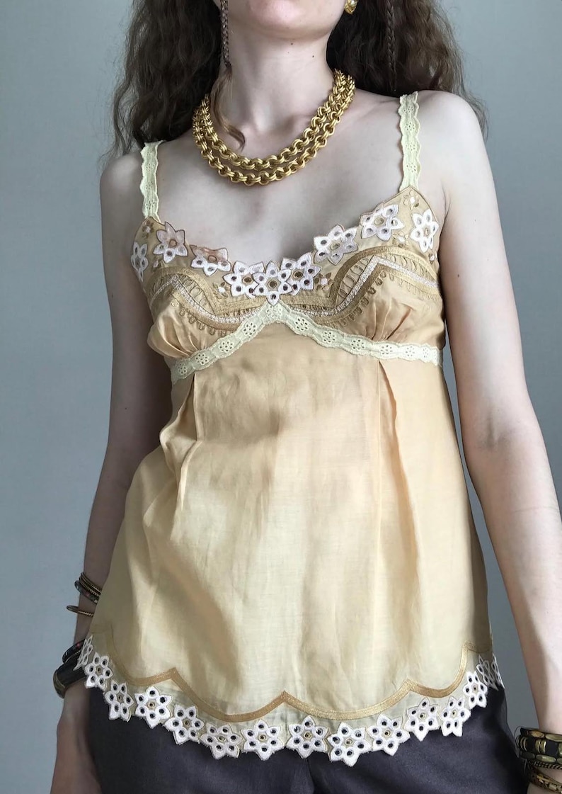 May include: A light yellow camisole with floral embroidery and lace trim. The top features delicate spaghetti straps and a scalloped hemline. A gold chain necklace is worn around the neck. The camisole is a vintage style.