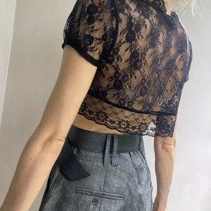 Pu&ograve; includere: Top corto in pizzo nero con maniche corte. Il top presenta un motivo floreale in pizzo e un orlo smerlato. Abbinato a pantaloni grigi a vita alta e una cintura nera.