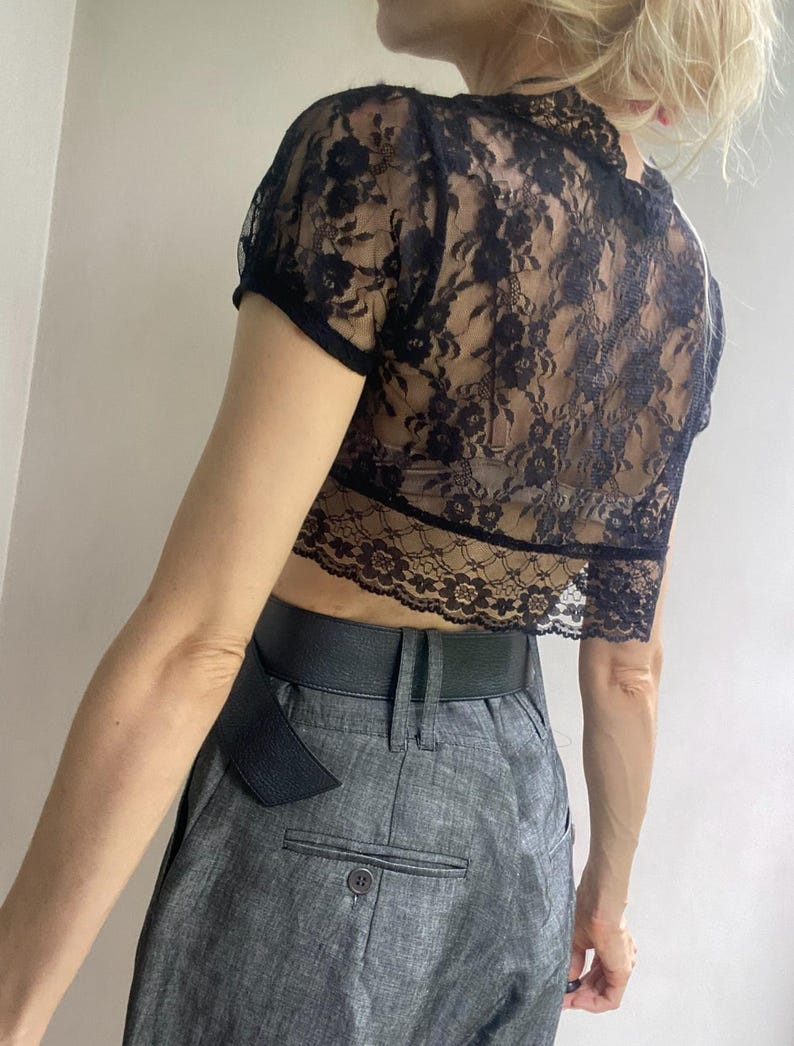 Pu&ograve; includere: Top corto in pizzo nero con maniche corte. Il top presenta un motivo floreale in pizzo e un orlo smerlato. Abbinato a pantaloni grigi a vita alta e una cintura nera.