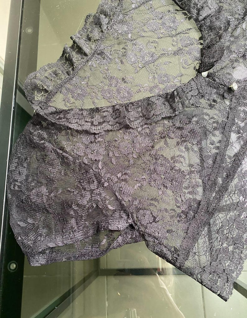 Pu&ograve; includere: Primo piano di un capo in pizzo nero con un motivo floreale. Il tessuto &egrave; semitrasparente, con un bordo a balze e piccoli bottoni perlati. Il capo sembra essere lingerie o un articolo delicato simile.