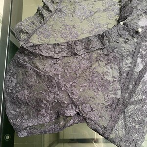 Pu&ograve; includere: Primo piano di un capo in pizzo nero con un motivo floreale. Il tessuto trasparente presenta un bordo a balze e piccoli bottoni perlati. Il capo sembra essere lingerie o un articolo delicato simile.