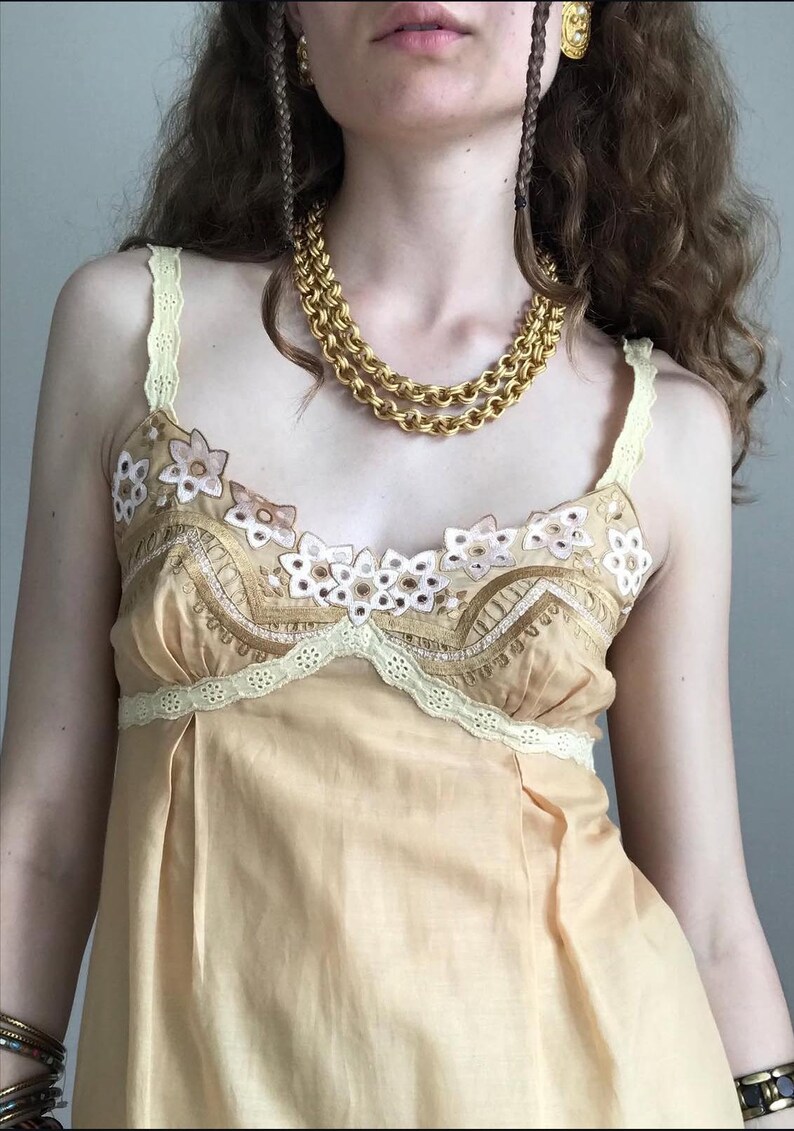 May include: A light peach-colored camisole top with floral embroidery and lace trim. The top features a scoop neckline and spaghetti straps. A gold chain necklace is worn over the top.