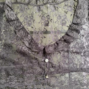 Pu&ograve; includere: Un top in pizzo grigio scuro trasparente con un motivo floreale. Il top presenta un colletto a balze, uno scollo a V e una chiusura con bottoni. Il tessuto delicato crea un look romantico ed elegante.
