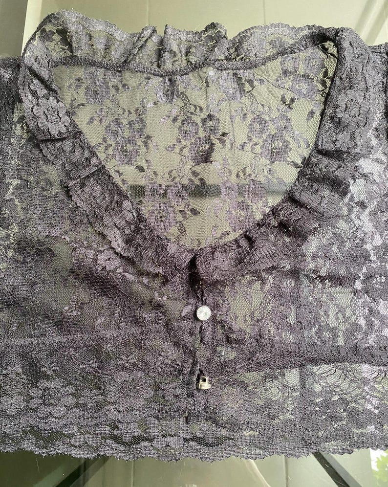Pu&ograve; includere: Un top in pizzo grigio scuro trasparente con un motivo floreale. Il top presenta un colletto a balze, uno scollo a V e una chiusura con bottoni. Il tessuto delicato crea un look romantico ed elegante.