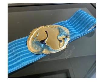 May include: A gold-toned belt buckle with a blue and gold design, attached to a blue elastic band. The buckle features a detailed, abstract design. The belt is displayed on a reflective surface.