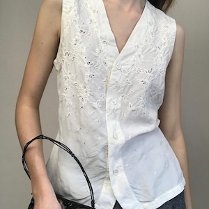 May include: White sleeveless vest with floral embroidery and button-up front. The vest has a V-neck and is made of a lightweight fabric. The vest is part of a vintage clothing collection.