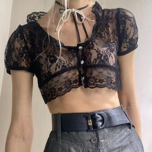 Pu&ograve; includere: Top corto in pizzo nero con maniche corte e scollatura smerlata. Il top presenta una chiusura con bottoni sul davanti ed &egrave; abbinato a una cintura nera e pantaloni di jeans grigi. Una collana girocollo di perle aggiunge un tocco delicato.
