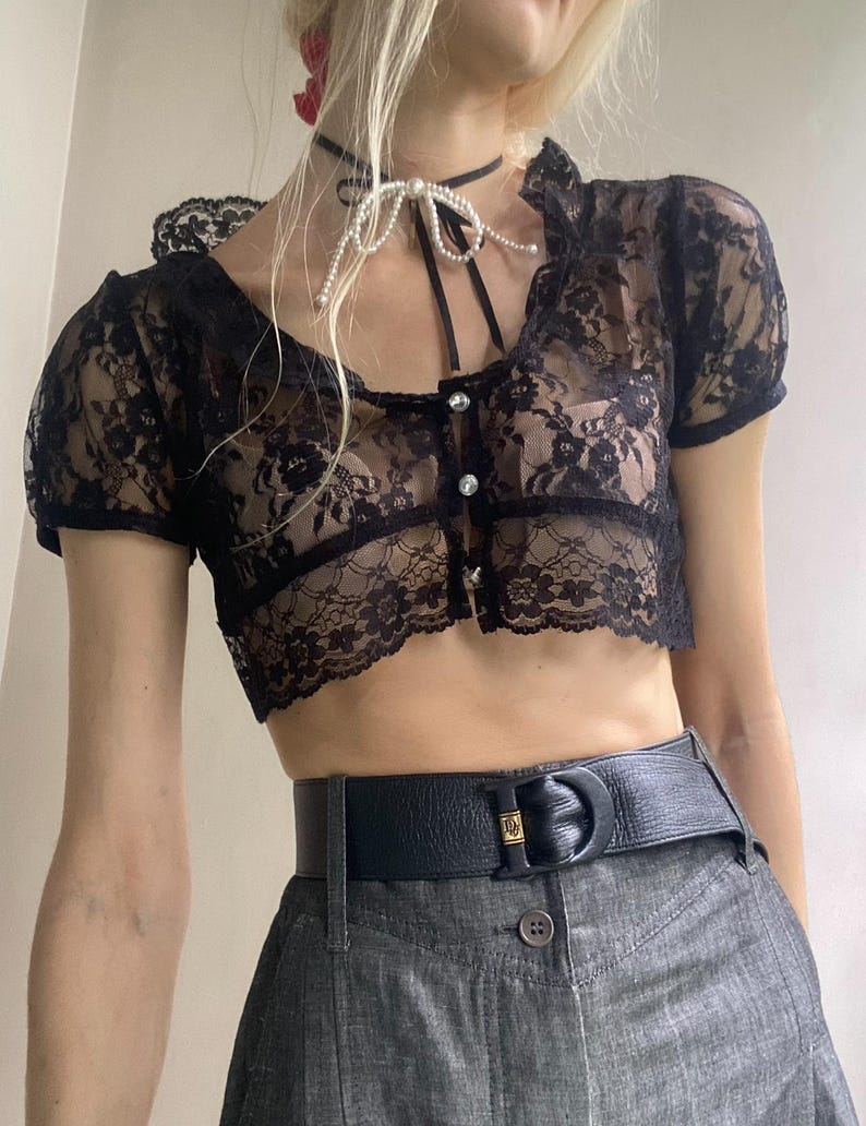 Pu&ograve; includere: Top corto in pizzo nero con maniche corte e scollatura smerlata. Il top presenta una chiusura con bottoni sul davanti ed &egrave; abbinato a una cintura nera e pantaloni di jeans grigi. Una collana girocollo di perle aggiunge un tocco delicato.