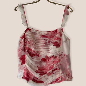 May include: A sleeveless top with a floral pattern in shades of red and white. The top features ruffled straps and a ruched bodice. The garment is hanging on a gold-colored hanger.
