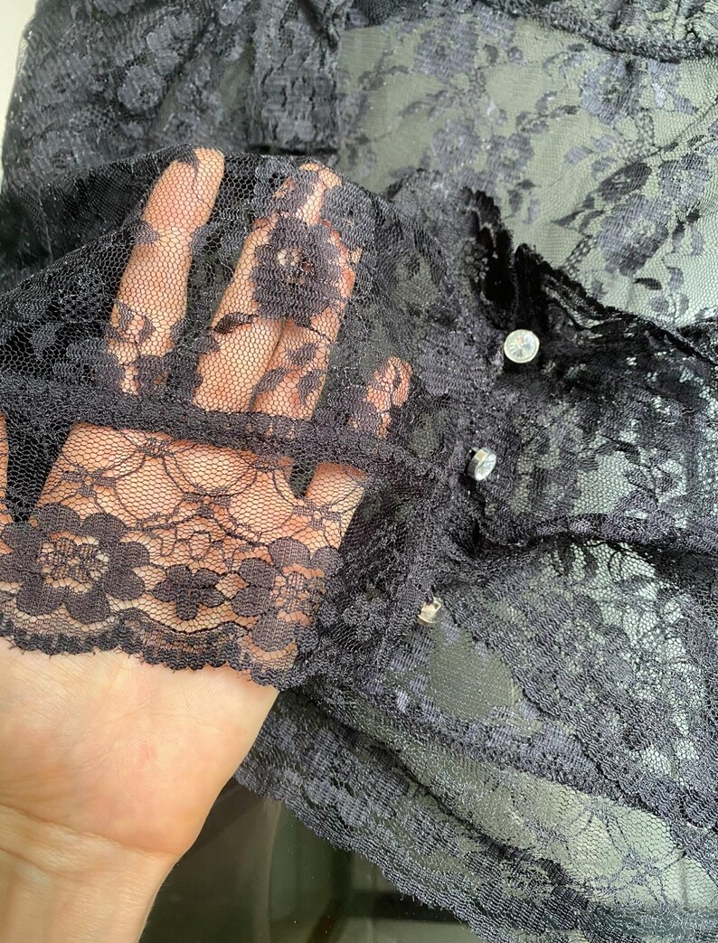 Pu&ograve; includere: Primo piano di un capo in pizzo nero. Il tessuto delicato presenta un motivo floreale ed &egrave; semitrasparente. Il capo ha bottoni decorativi. Il pizzo &egrave; tenuto in alto, mostrando i dettagli e la consistenza del materiale.