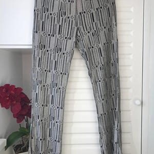 May include: Wide-leg trousers in a grey and black pattern. The trousers feature a black sequined waistband and are suspended from a decorative gold hanger. The design incorporates vertical lines and geometric shapes.