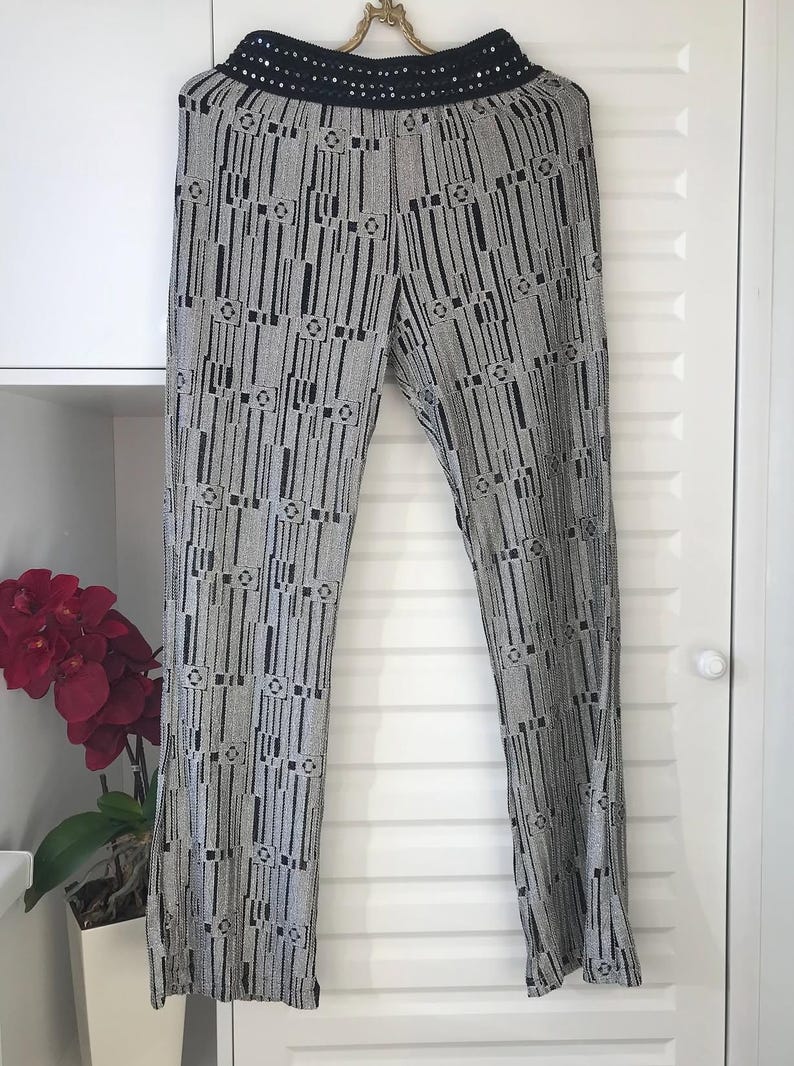 May include: Wide-leg trousers in a grey and black pattern. The trousers feature a black sequined waistband and are suspended from a decorative gold hanger. The design incorporates vertical lines and geometric shapes.