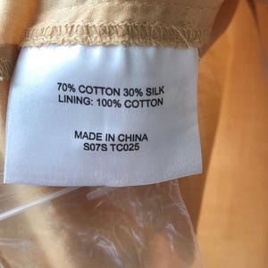 May include: A close-up of a label with text indicating the fabric composition: 70% cotton, 30% silk, and a 100% cotton lining. The label also states "Made in China" and includes the code "S07S TC025". The label is attached to a peach-colored fabric.