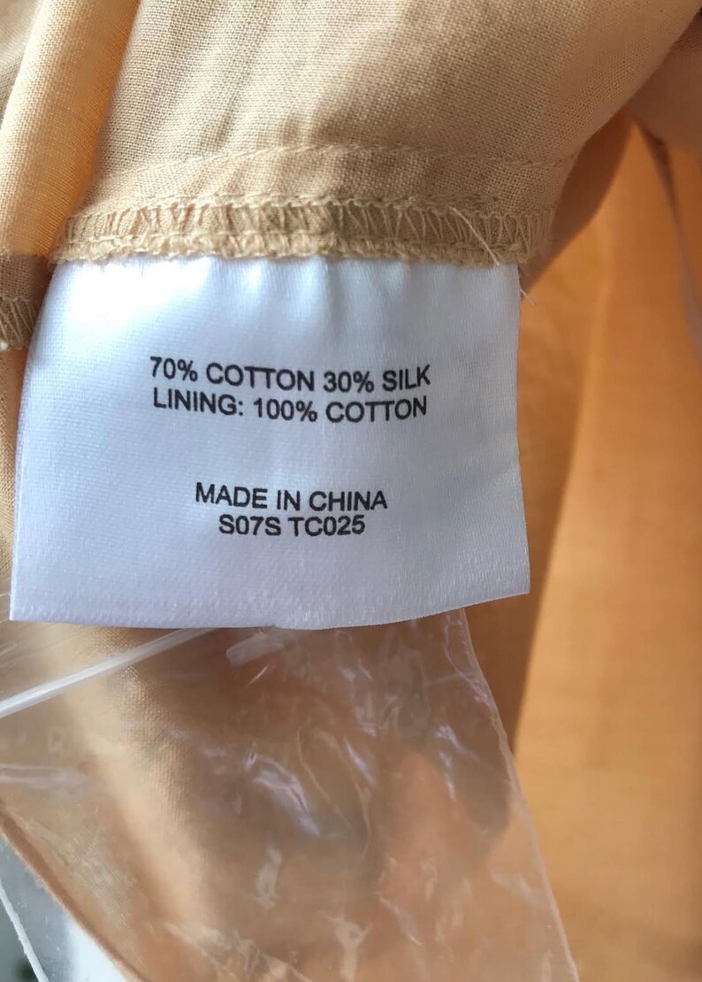 May include: A close-up of a label with text indicating the fabric composition: 70% cotton, 30% silk, and a 100% cotton lining. The label also states "Made in China" and includes the code "S07S TC025". The label is attached to a peach-colored fabric.