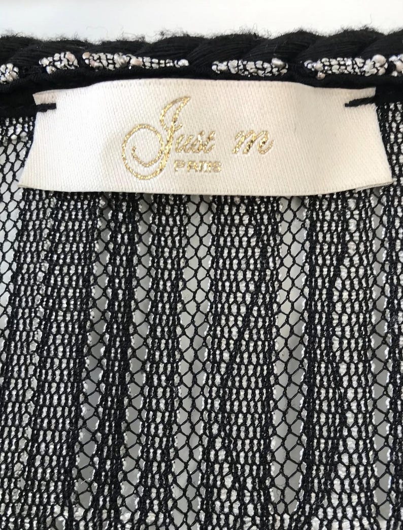May include: Close-up of a black lace garment with a white label. The label features gold lettering that reads "Just m PARIS". The top edge has a black trim with silver accents.