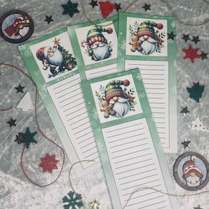 May include: A collection of Christmas-themed notepads featuring illustrations of Santa Claus and gnomes. The notepads have a green border and lined pages. Decorative wooden ornaments and star-shaped confetti surround the notepads, creating a festive holiday scene.