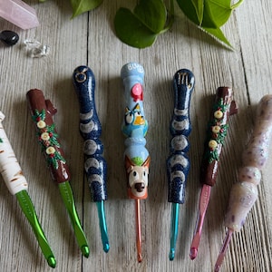 May include: A set of eight colorful crochet hooks with different designs. The hooks feature a variety of themes, including zodiac signs, animals, and nature. The hooks are made of wood and have a smooth, polished finish.
