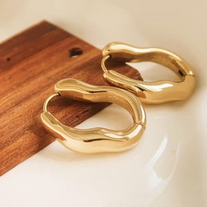 May include: A pair of gold-colored hoop earrings with a unique, organic shape. The earrings are smooth and shiny, reflecting light. They are resting on a wooden surface and a white surface.