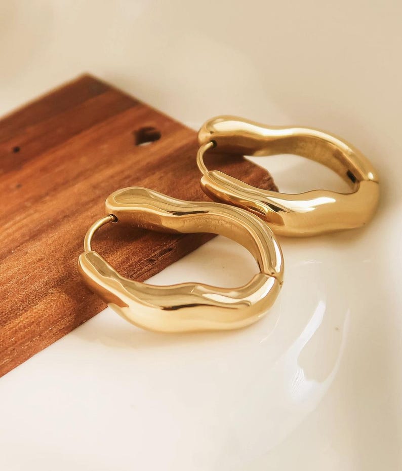May include: A pair of gold-colored hoop earrings with a unique, organic shape. The earrings are smooth and shiny, reflecting light. They are resting on a wooden surface and a white surface.