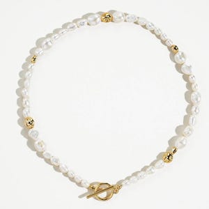 May include: A white pearl necklace with gold accents. The necklace features a variety of irregularly shaped pearls interspersed with small gold beads. The clasp is a gold toggle closure. The necklace is displayed on a white background.
