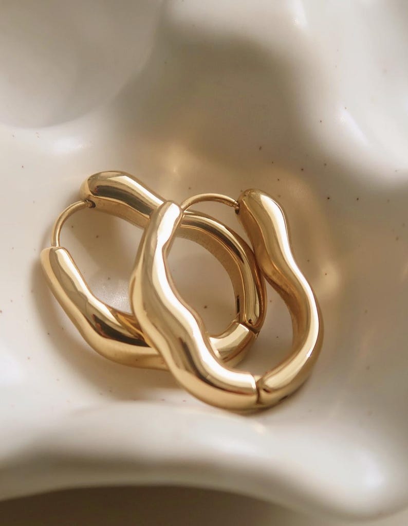 May include: A pair of gold-colored hoop earrings with a unique, organic shape. The earrings have a smooth, polished finish and are displayed on a white, slightly textured surface. The design is modern and minimalist, suitable for everyday wear.