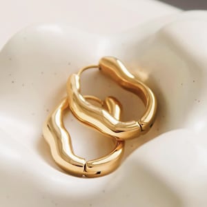 May include: A pair of gold-colored hoop earrings with a unique, wavy design. The earrings have a smooth, polished finish and a secure clasp closure. The hoops are displayed on a white, textured surface.