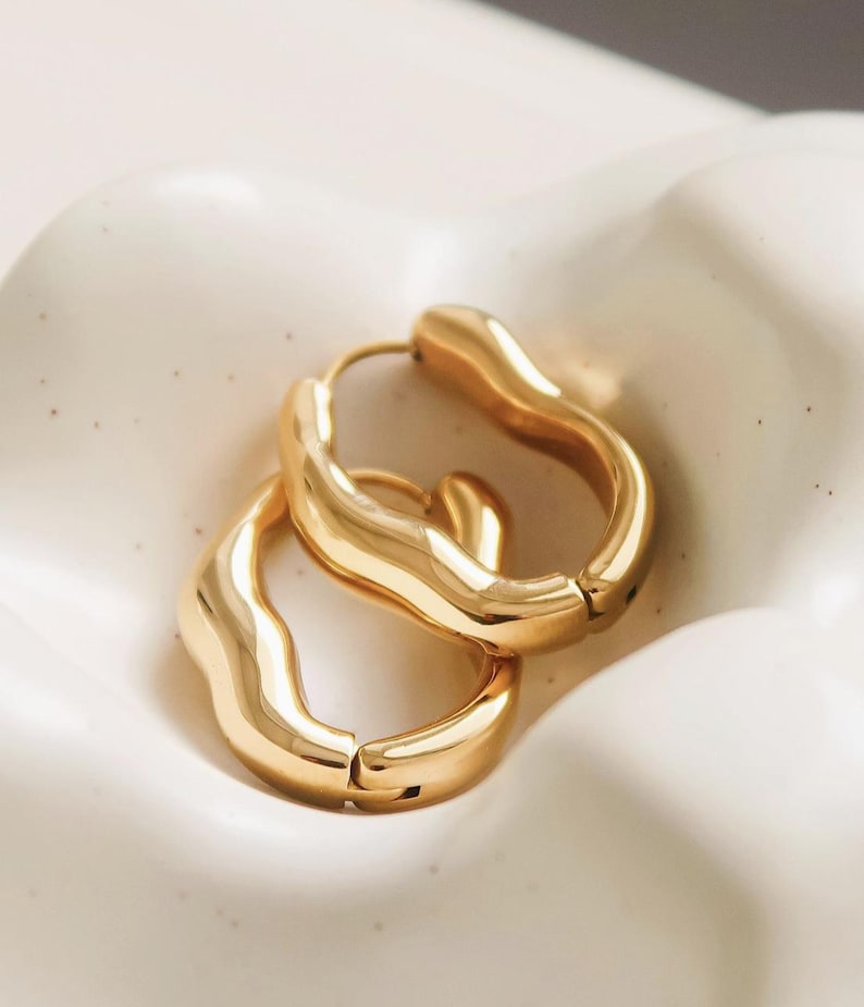 May include: A pair of gold-colored hoop earrings with a unique, wavy design. The earrings have a smooth, polished finish and a secure clasp closure. The hoops are displayed on a white, textured surface.