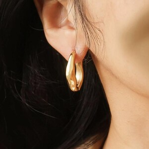 May include: A close-up shot of a gold-colored hoop earring. The earring has a thick, rounded design with a smooth, polished surface. The earring is worn on an ear, with the skin and hair visible in the background.