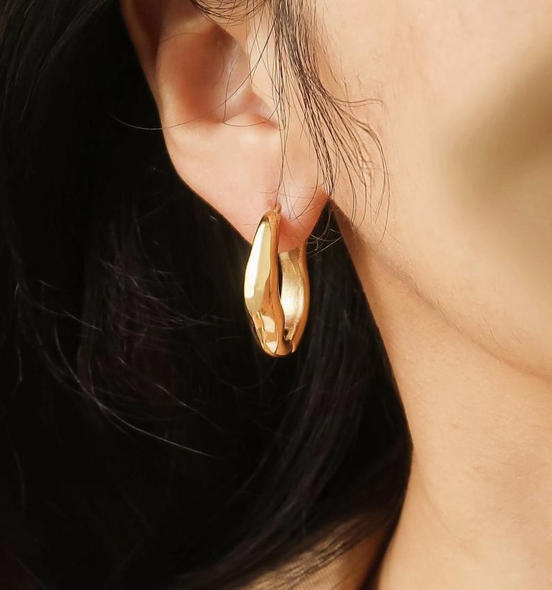 May include: A close-up shot of a gold-colored hoop earring. The earring has a thick, rounded design with a smooth, polished surface. The earring is worn on an ear, with the skin and hair visible in the background.