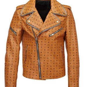 Op de afbeelding: Een bruinleren jas met een herhalend zwart diamant- en logopatroon. De jas heeft een reverskraag, asymmetrische ritssluiting en meerdere zakken met ritssluiting. De mouwen hebben ritsen bij de manchetten. De jas is een klassieke bikerstijl.