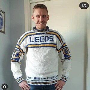 May include: A cream-coloured knitted jumper with blue and yellow detailing. The word "LEEDS" is across the chest. The sleeves feature a crest and the words "MARCHING ON TOGETHER" are at the bottom. The jumper is worn by a person.