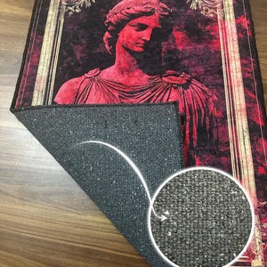 May include: A red and gold framed rug with a bust of a woman in a red toga. The rug has a cotton base.